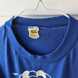 May include: A vintage blue t-shirt with a crew neck and a distressed collar. The shirt has a tag that reads "Velva Sheen". The shirt is hanging on a white metal hanger.