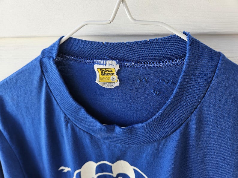 May include: A vintage blue t-shirt with a crew neck and a distressed collar. The shirt has a tag that reads "Velva Sheen". The shirt is hanging on a white metal hanger.