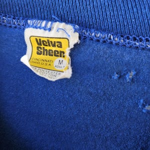 May include: Close-up of a blue t-shirt collar with a tag. The tag is yellow and white with the text "Velva Sheen" and "Cincinnati, Ohio, U.S.A." The tag also indicates the size is "M" for adult.