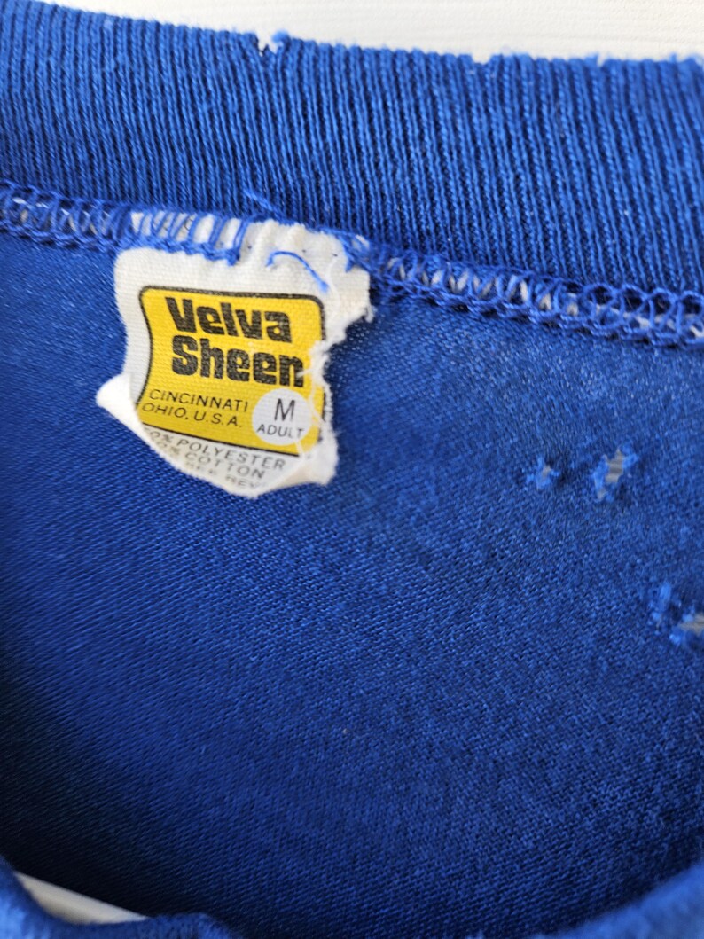 May include: Close-up of a blue t-shirt collar with a tag. The tag is yellow and white with the text "Velva Sheen" and "Cincinnati, Ohio, U.S.A." The tag also indicates the size is "M" for adult.