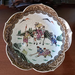 May include: A white ceramic bowl with a brown and gold floral design. The bowl features a scene of people riding horses and a goat in a mountainous landscape.