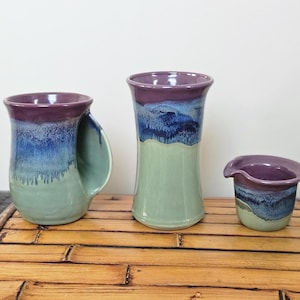 May include: Three handmade ceramic pieces: a mug with a large handle, a tall vase, and a small creamer. Each piece features a color gradient of purple, blue, and green. The mug is on the left, the vase in the middle, and the creamer on the right.