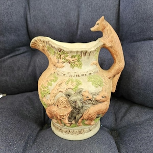 Puede incluir: Jarra de cerámica con asa en forma de zorro y una escena en relieve detallada de zorros y otros animales. La jarra es principalmente de color beige con detalles verdes y marrones, y el asa es de color marrón claro. La base es de color beige claro.