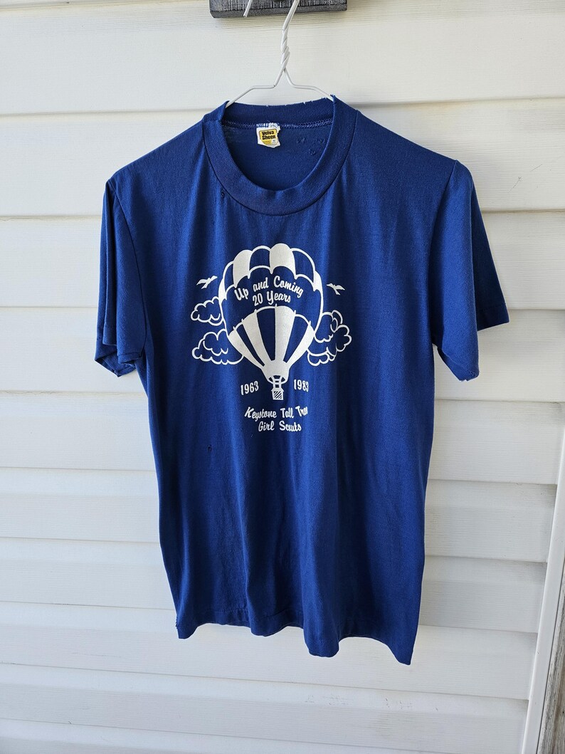 May include: A navy blue t-shirt with a white graphic of a hot air balloon, clouds, and text that reads "Up and Coming 20 Years" and "1963-1983 Keystone Tall Tree Girl Scouts."
