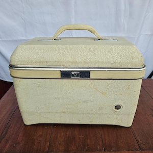 Puede incluir: Maleta de tocador vintage de color crema con exterior texturizado y asa curva. La maleta presenta un ribete metálico plateado y un cierre seguro. Un pequeño botón redondo es visible en la parte delantera. La maleta está sobre una superficie de madera.