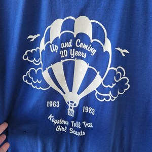 May include: A blue t-shirt with a white hot air balloon graphic. The graphic includes the text "Up and Coming 20 Years", "1963", "1983", and "Keystone Tall Tree Girl Scouts". The shirt has a crew neck.