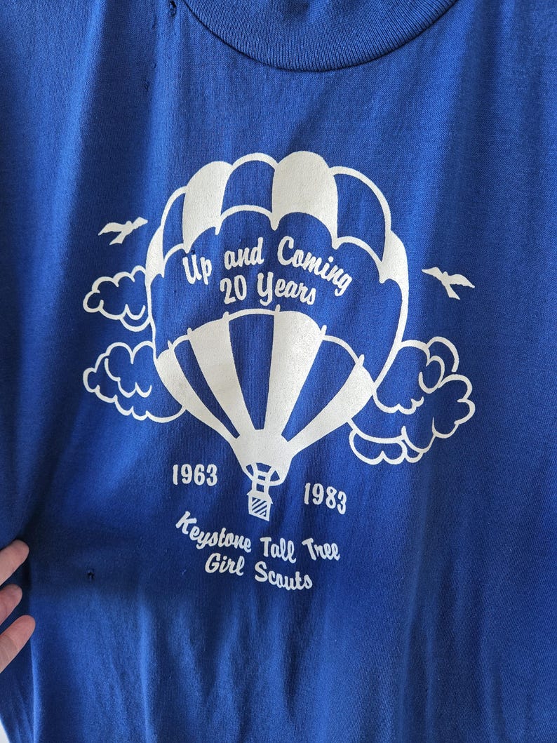 May include: A blue t-shirt with a white hot air balloon graphic. The graphic includes the text "Up and Coming 20 Years", "1963", "1983", and "Keystone Tall Tree Girl Scouts". The shirt has a crew neck.