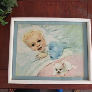May include: A framed watercolor painting of a baby sleeping in a white crib with a white kitten. The baby is wearing a blue and white dress and has blonde hair. The kitten is lying on a pink blanket.