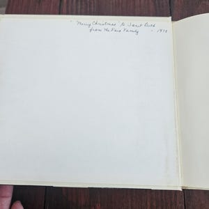 Op de afbeelding: Een open boek met een witte pagina en handgeschreven tekst die luidt: "Merry Christmas to Janet Beck from the Fairs Family - 1978". Het boek ligt op een donker houten oppervlak.