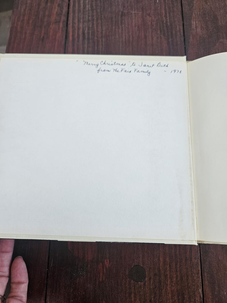Op de afbeelding: Een open boek met een witte pagina en handgeschreven tekst die luidt: "Merry Christmas to Janet Beck from the Fairs Family - 1978". Het boek ligt op een donker houten oppervlak.