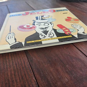 Op de afbeelding: Vintage boek getiteld "Amazing Jello Recipes" met een stripfiguur tovenaar op de cover. De cover heeft een cr&egrave;mekleurige achtergrond met zwart-wit illustraties en de tekst "PLUS: Magic Tricks By Marvello The Great".