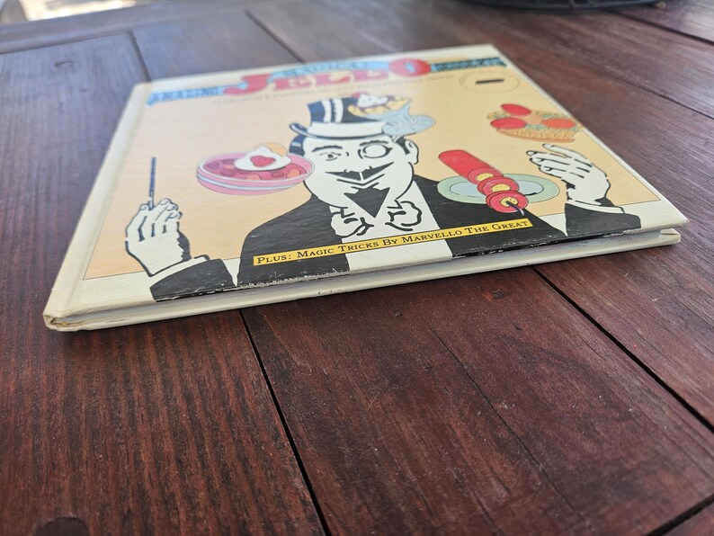 Op de afbeelding: Vintage boek getiteld "Amazing Jello Recipes" met een stripfiguur tovenaar op de cover. De cover heeft een cr&egrave;mekleurige achtergrond met zwart-wit illustraties en de tekst "PLUS: Magic Tricks By Marvello The Great".
