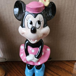 May include: A ceramic figurine of Minnie Mouse wearing a pink dress with white polka dots, a pink hat with a yellow flower, and blue shoes.