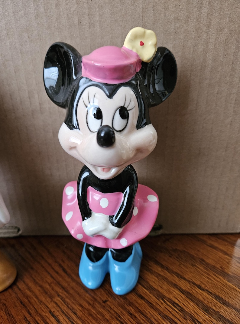 May include: A ceramic figurine of Minnie Mouse wearing a pink dress with white polka dots, a pink hat with a yellow flower, and blue shoes.