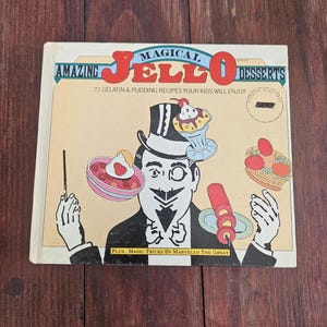 Op de afbeelding: Een vintage kookboek getiteld "Amazing Jello Desserts" met een cartoon illustratie van een goochelaar. De cover bevat de tekst "Magical Jello Desserts" en "72 Gelatin & Pudding Recipes Your Kids Will Enjoy."