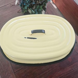 May include: An oval-shaped roasting pan with a pale yellow lid featuring a dark gray handle and a small dark gray carrot graphic. The pan has a dark gray base and a ribbed texture on the lid, showing signs of wear on the painted surface.