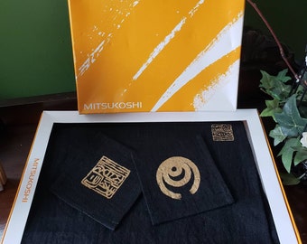 May include: A black textile set with gold designs, presented in a white and gold box. The set includes a rectangular mat, two smaller square pieces, and a small square with a design. The box has the word "MITSUKOSHI" printed on it.