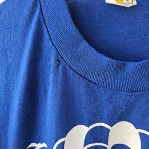 May include: Close-up of a vintage blue t-shirt with a white graphic design. The graphic includes the words "Up and Coming" and "20 Years". The shirt has a crew neck and a tag that reads "Velva Sheen".