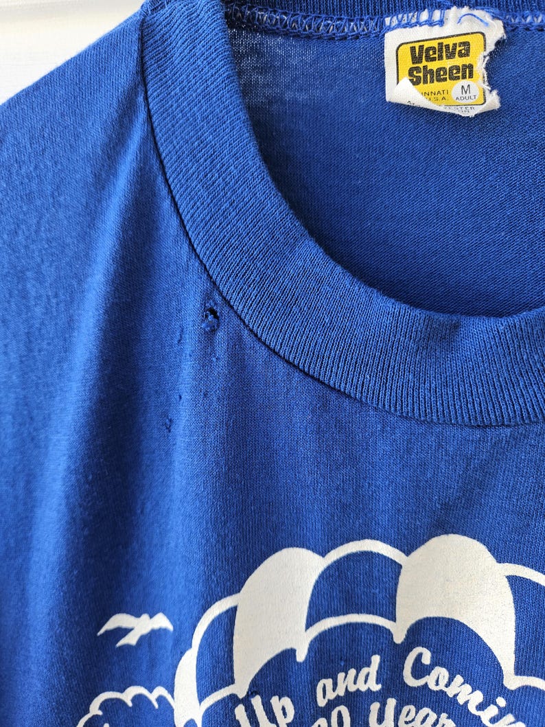 May include: Close-up of a vintage blue t-shirt with a white graphic design. The graphic includes the words "Up and Coming" and "20 Years". The shirt has a crew neck and a tag that reads "Velva Sheen".