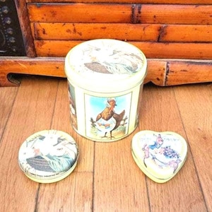 May include: A cylindrical cream-colored tin with a lid, featuring a central illustration of a hen wearing a bonnet and apron. Beside it are two smaller tins: one round with a lid depicting a bird, and one heart-shaped with a floral design.