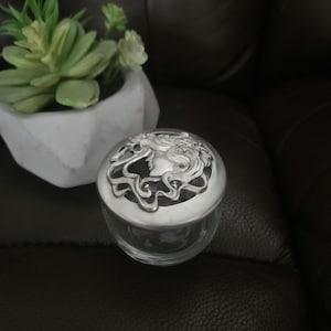 Pode incluir: Um frasco de vidro transparente com uma tampa decorativa prateada. A tampa apresenta um perfil detalhado do rosto de uma mulher com cabelo esvoaçante. O frasco está sobre uma superfície escura, com um vaso geométrico branco e uma planta suculenta ao fundo.