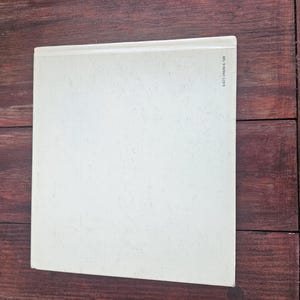 Op de afbeelding: Een boek met een lichtcr&egrave;mekleurige kaft, tegen een donkere houtnerf achtergrond. De kaft van het boek is effen, met een licht getextureerd oppervlak. De rug is zichtbaar en een reeks cijfers is aan de rechterkant gedrukt.