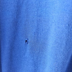 May include: Close-up of a textured, bright blue fabric with a small hole. The fabric appears to be a knit or woven material, with visible fibers and a slightly worn appearance. The hole is located near the center of the image.