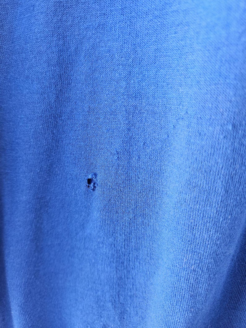 May include: Close-up of a textured, bright blue fabric with a small hole. The fabric appears to be a knit or woven material, with visible fibers and a slightly worn appearance. The hole is located near the center of the image.