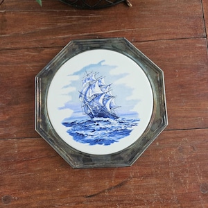 Puede incluir: Un plato decorativo octogonal con marco plateado. El plato presenta una detallada ilustración en azul y blanco de un gran velero en el mar. El barco está representado con velas ondeantes y rodeado de olas.
