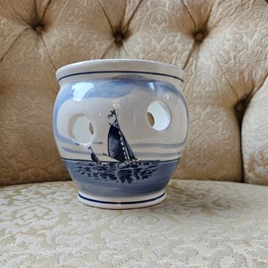 May include: A white ceramic planter with a blue and white Delft design of sailing ships. The planter has three holes in the side for drainage.