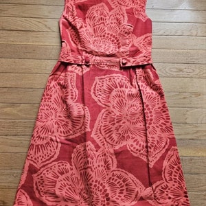 May include: A sleeveless red dress with a floral print. The dress has a fitted bodice and a full skirt. The flowers are a lighter shade of red and have a delicate, intricate design.