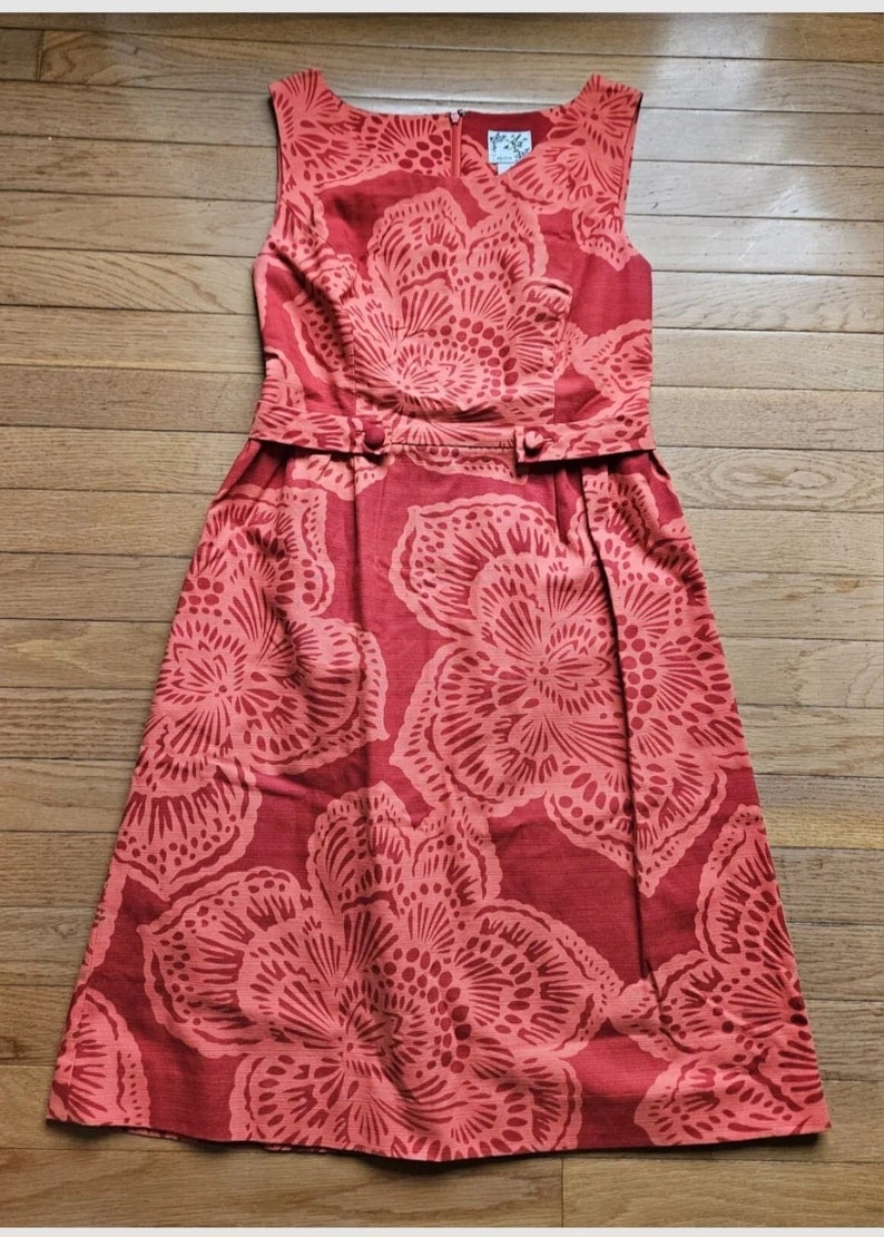 May include: A sleeveless red dress with a floral print. The dress has a fitted bodice and a full skirt. The flowers are a lighter shade of red and have a delicate, intricate design.