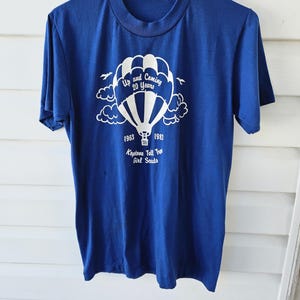 May include: A vintage blue t-shirt with a white graphic of a hot air balloon, clouds, and text that reads "Up and Coming 20 Years" and "1963-1983 Keystone Tall Tree Girl Scouts". The shirt has short sleeves and a crew neck.