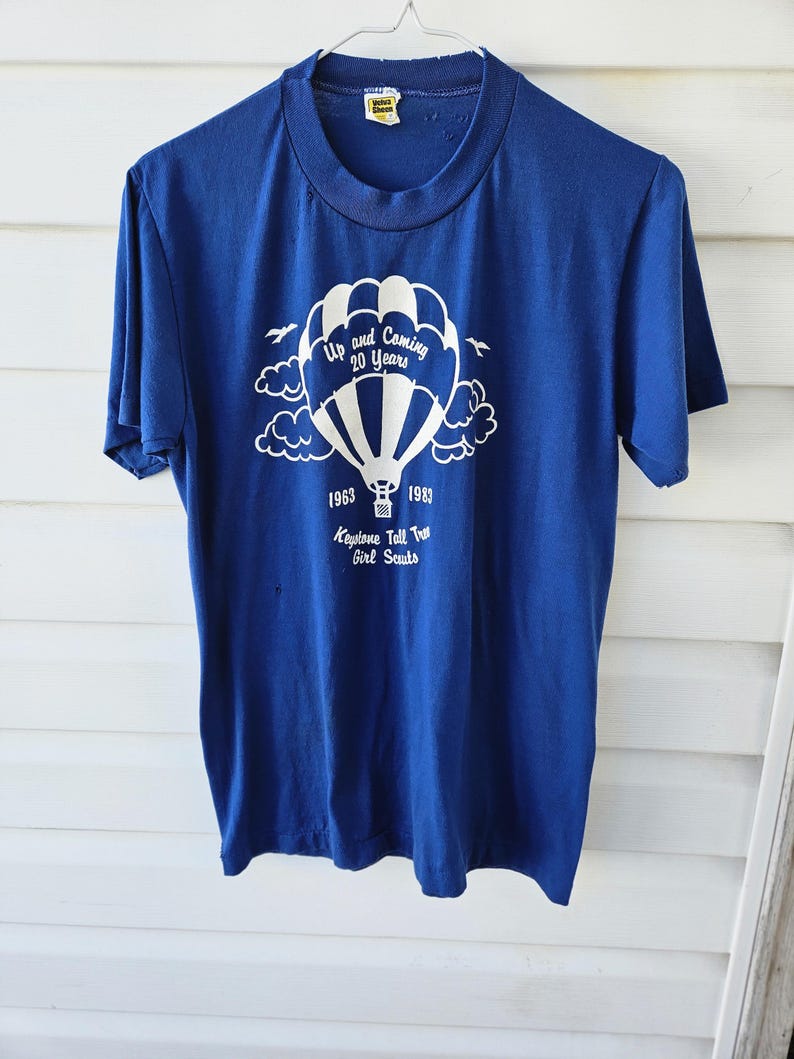 May include: A vintage blue t-shirt with a white graphic of a hot air balloon, clouds, and text that reads "Up and Coming 20 Years" and "1963-1983 Keystone Tall Tree Girl Scouts". The shirt has short sleeves and a crew neck.