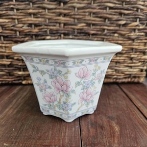 May include: A white ceramic planter with a square rim and a tapered body. The exterior features a delicate floral pattern with pink magnolia blossoms, small blue flowers, and green leaves, accented by a thin decorative border along the top edge.