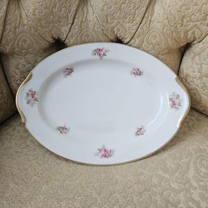 May include: Oval white porcelain serving platter with gold trim and pink floral details. The platter has a raised edge and handles. A classic piece for serving food. The platter is approximately 30 cm in length.