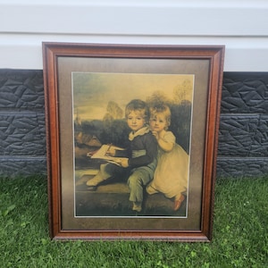 May include: A framed print featuring two children, a boy and a girl, in a classic style. The boy is seated, holding a pencil and paper, while the girl stands beside him. The frame is a dark brown color, and the artwork has a vintage aesthetic.