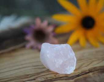 May include: A pale pink rose quartz crystal with a rough, natural texture. The crystal is sitting on a wooden surface.