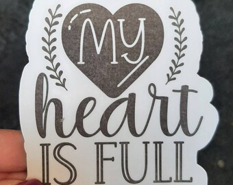 May include: A white sticker with a heart-shaped design. The heart is filled with a dark gray color and the words "MY" are written in white. Below the heart, the words "heart IS FULL" are written in a script font. The sticker has a scalloped edge.