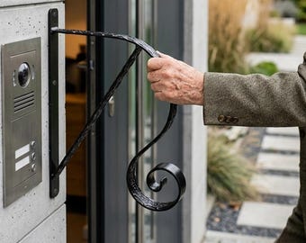 Può includere: Un corrimano in ferro battuto nero con un design a volute decorative è montato su un muro di cemento accanto a una porta moderna. Il corrimano è collegato a un sistema di citofono a parete.