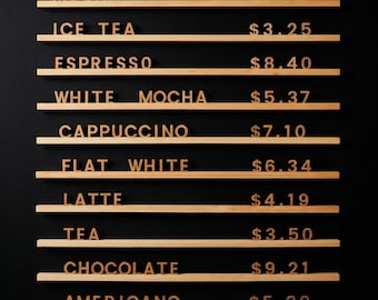 May include: A wall-mounted menu board featuring ten horizontal light wood shelves. Each shelf displays individual wooden letters and numbers spelling out beverage names and prices: MOCHA $9.40, ICE TEA $3.25, ESPRESSO $8.40, WHITE MOCHA $5.37, CAPPUCCINO $7.10, FLAT WHITE $6.34, LATTE $4.19, TEA $3.50, CHOCOLATE $9.21, and AMERICANO $5.30.