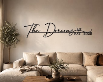 May include: Black metal wall art with the text "The Dorions" and "EST 2020" in a cursive font. The sign is mounted on a neutral-colored wall above a beige sectional sofa. A decorative olive tree is in the background.