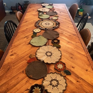 May include: A wooden dining table with a chevron pattern, decorated with a collection of handmade crochet placemats and decorative pieces in various earth tones. The placemats are round and flower-shaped, adding a rustic, handcrafted aesthetic.