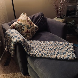 Op de afbeelding: Een donkergrijze chaise longue met een gepatroneerd kussen en een blauwe en witte gehaakte deken.