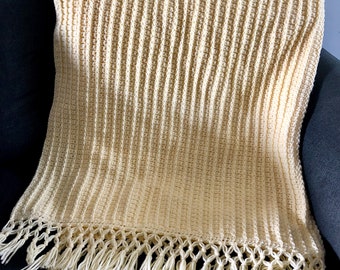 May include: A crocheted throw blanket in a cream colour with a fringe border. The blanket is folded and placed on a dark grey sofa.