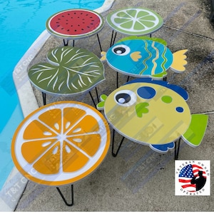 May include: Five colorful, wooden side tables with different designs. The tables feature a watermelon, a lime, a green leaf, a blue fish, and a yellow fish. Each table has a black metal base with three legs.