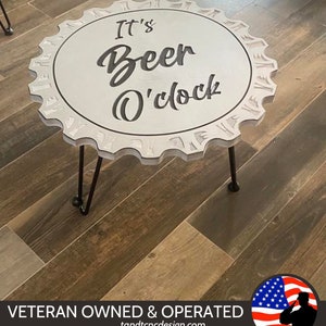 May include: White wooden table shaped like a bottle cap with the text "It's Beer O'clock" on top. The table has two black hairpin legs. The table is on a wood floor.
