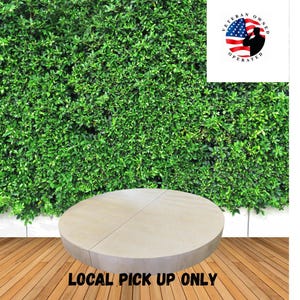 Op de afbeelding: Een ronde, lichtgeleurde stenen tafel met een tweedelig ontwerp, geplaatst op een houten terras. De achtergrond toont een dichte groene haag. De afbeelding bevat de tekst "LOCAL PICK UP ONLY" en een logo "VETERAN OWNED OPERATED".