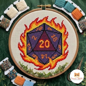 May include: A cross-stitch embroidery in a circular wooden hoop featuring a purple twenty-sided die surrounded by stylised orange and yellow flames. The die displays the number 20 in the centre, with other numbers visible on its triangular faces.