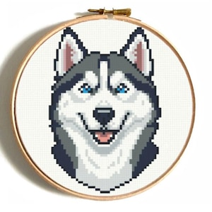 May include: A cross-stitch embroidery of a smiling Siberian Husky dog. The dog has blue eyes, gray and white fur, and is framed in a wooden embroidery hoop. The background is white.